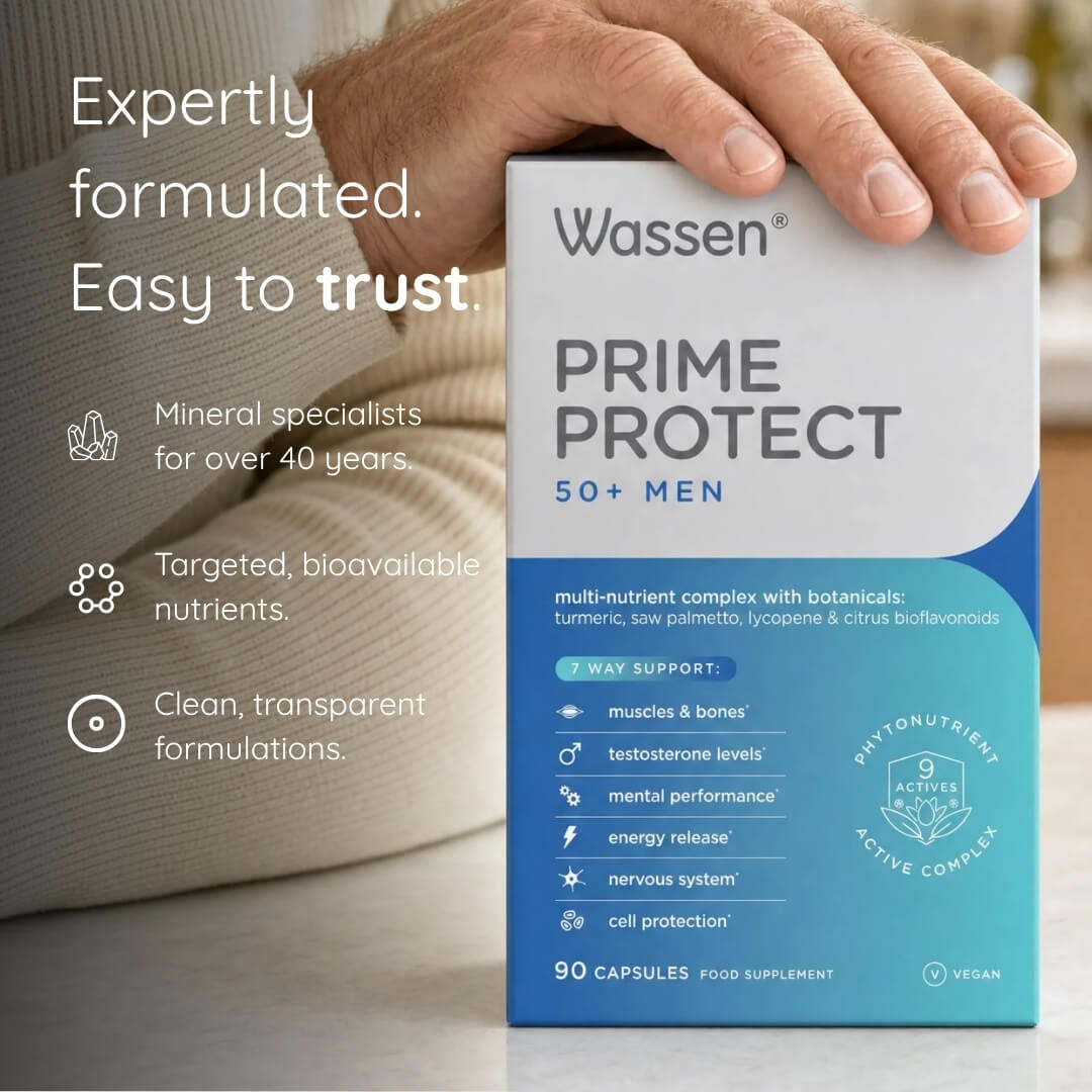 A hand holds a box of Wassen Prime Protect 50+ Men multivitamin capsules. The packaging is white and blue, and the background shows a blurred indoor setting with a man's arm and a beige sofa.