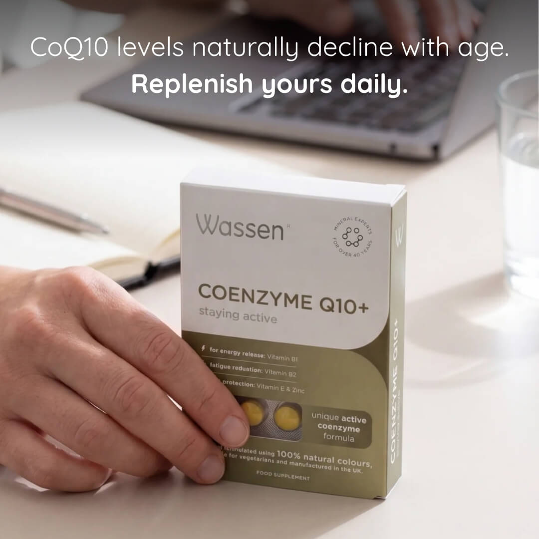 A hand holds a box of Wassen Coenzyme Q10+ supplements on a white desk, with a blurred laptop and glass of water in the background. Text at the top reads, 'CoQ10 levels naturally decline with age.