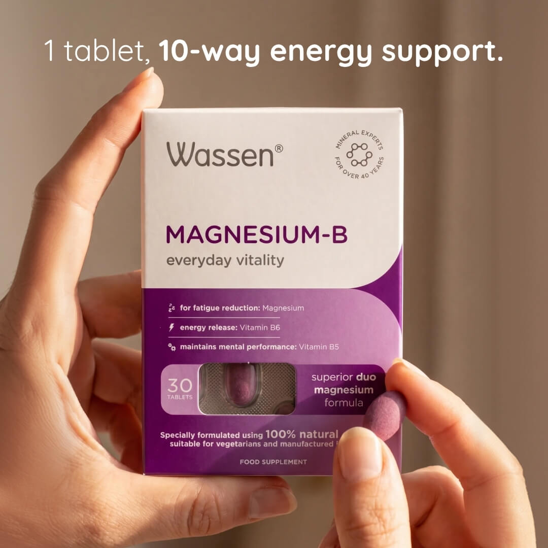 A person's hands holding a box of Wassen Magnesium-B dietary supplement. The white and purple box features text highlighting '1 tablet, 10-way energy support.