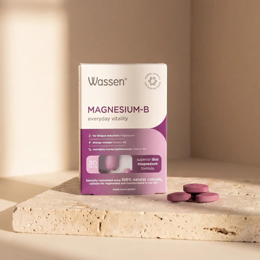 Wassen Magnesium-B supplement box standing on a light stone slab with three pinkish-purple tablets beside it, in a warm beige studio setting with soft shadows.