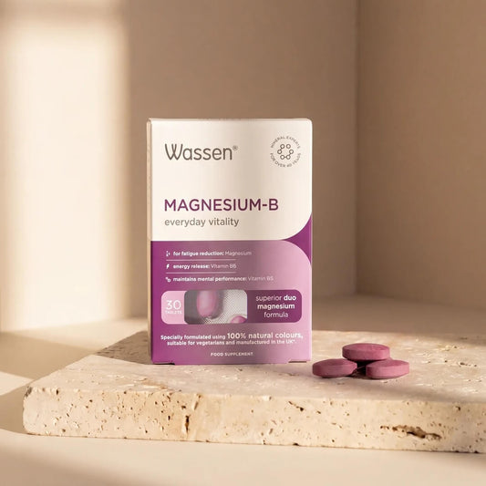 Wassen Magnesium-B supplement box standing on a light stone slab with three pinkish-purple tablets beside it, in a warm beige studio setting with soft shadows.