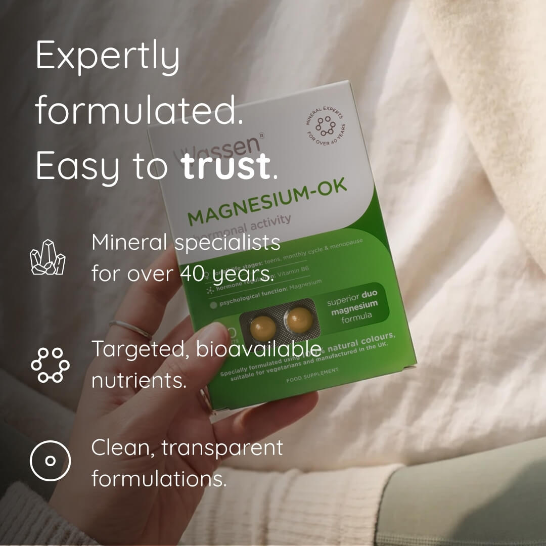 A hand holds a box of Tussin Magnesium-OK supplement in a cozy indoor setting with a cream-colored blanket. The packaging is white and green with two visible capsules inside.