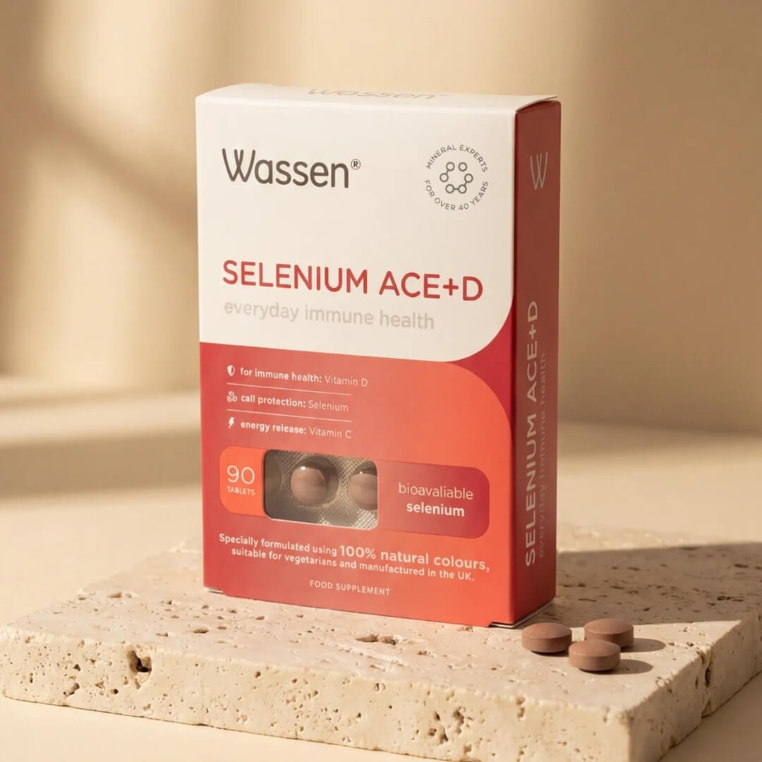 Wassen Selenium ACE+D supplement box in a warm beige and orange-red color scheme, styled on a textured stone slab with three brown capsules in the foreground.