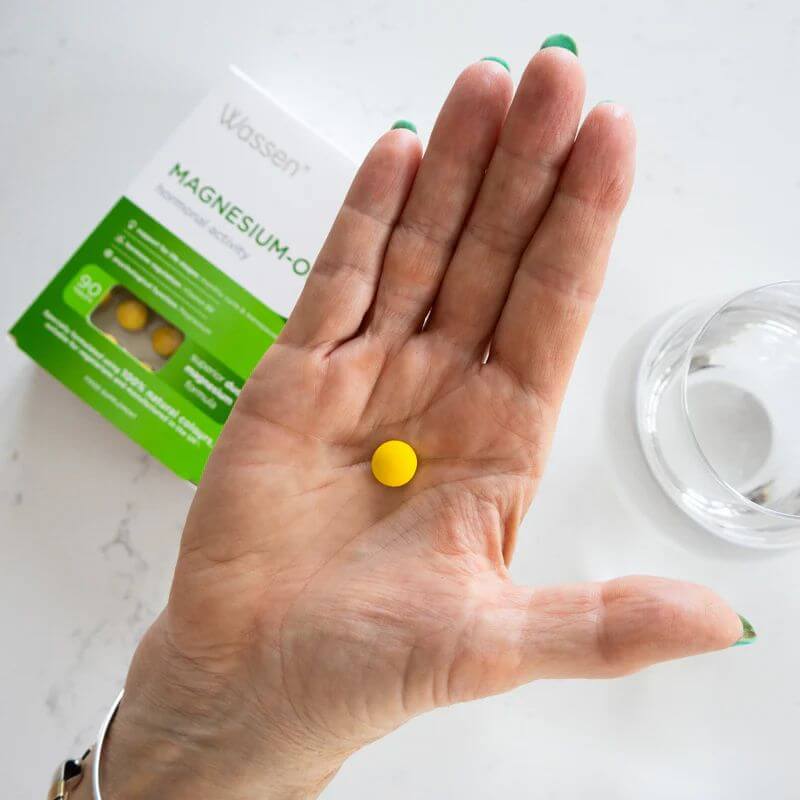 Wassen Magnesium-OK supplement in a woman's hand next to glass of water and pack of the supplement